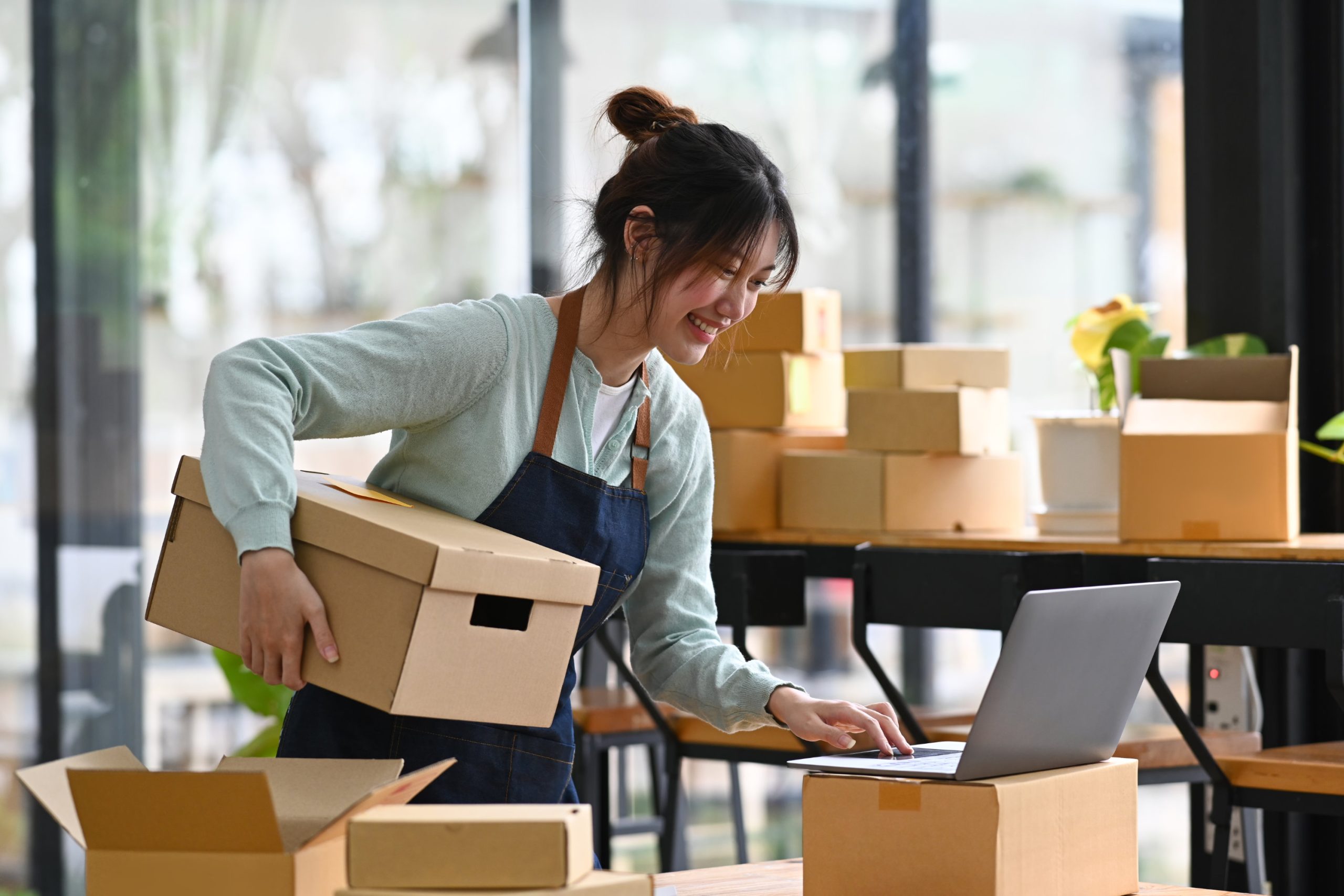 A woman holding a box in one hand and using a laptop with the other.