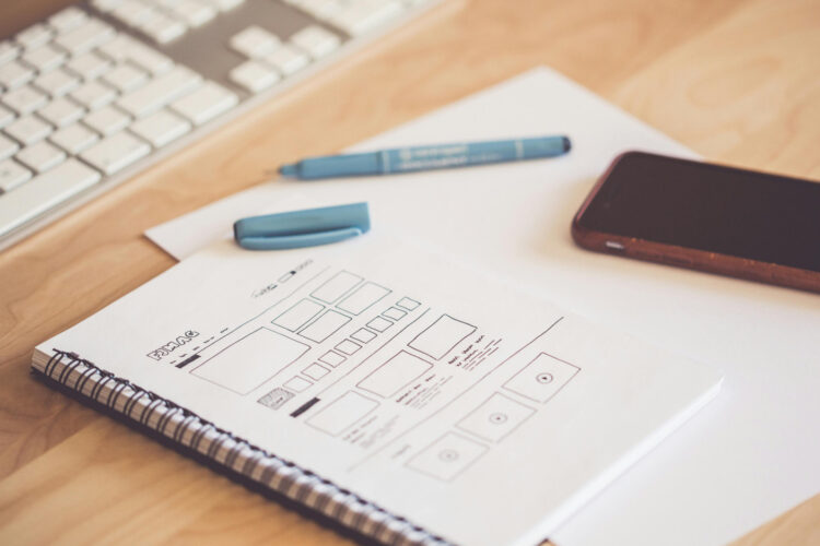 A notebook with a pen resting on it, showing sketched out wireframe designs for a website.