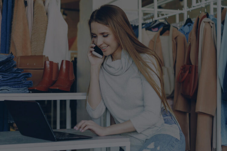 A woman on a phone in her clothes shop while sat at a laptop